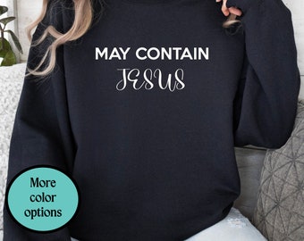 May include: Black sweatshirt with the text "MAY CONTAIN JESUS" in white block letters and cursive script. A teal circle with the words "More color options" is in the lower left corner. The sweatshirt is worn by a person.