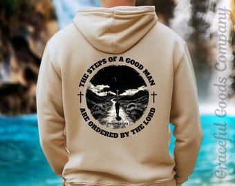 May include: Beige hoodie with a circular graphic on the back featuring a person walking on a path towards a cross under a night sky. The text reads "THE STEPS OF A GOOD MAN ARE ORDERED BY THE LORD" and "PSALMS 37:23".