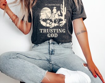 Könnte beinhalten: Dunkelgraues T-Shirt mit einer Wüstenlandschaft, die einen Cowboy auf einem Pferd, einen Kaktus und den Text "I'M JUST OUT HERE TRUSTING GOD" zeigt. Das Shirt wird mit hellblauen Jeans und weißen Turnschuhen getragen.