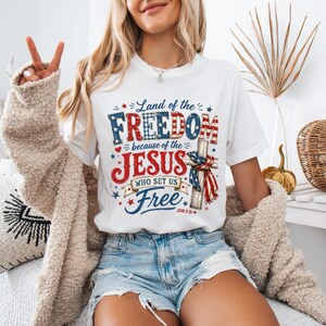 Könnte beinhalten: Weißes T-Shirt mit patriotischem Design. Die Grafik enthält die Worte "Land of the Freedom" und "Jesus Who Set Us Free" in Rot, Weiß und Blau. Das Design enthält Sterne und ein Kreuz.