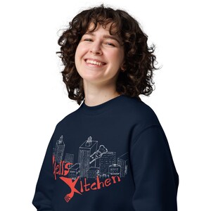 Könnte beinhalten: Ein marineblauer Pullover mit einem weißen Grafikdruck eines Stadtbildes und dem Text "Hell's Kitchen NYC" in Rot.