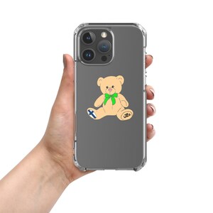 May include: Clear phone case with a cartoon teddy bear design. The bear is tan with a green bow, a Finnish flag, and the initials "JB". The phone case is held in a hand.