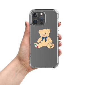 May include: Clear phone case featuring a tan teddy bear illustration with a blue bow tie and a Mexican flag on its left leg. The bear also has a logo on its right leg. The phone case is held by a person.