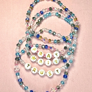May include: Five beaded bracelets with the words "Pray", "Wait", and "Trust" spelled out in gold letters on white beads. The bracelets are made with clear, blue, and pink beads.