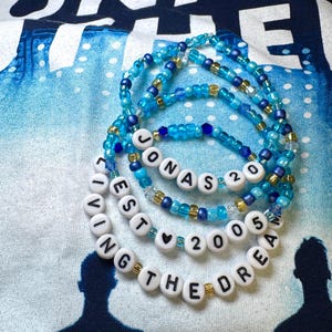 May include: A blue and gold beaded necklace with white letter beads spelling "Jonas Brothers Est 2005 Living The Dream" with a heart charm.