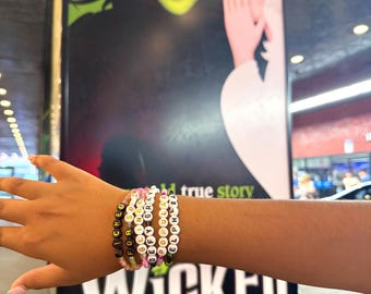 May include: Several beaded bracelets spelling out words like "WICKED", "FOREVER", and character names. The bracelets are in various colors, including black, pink, and white. The background features a poster for the musical "Wicked".