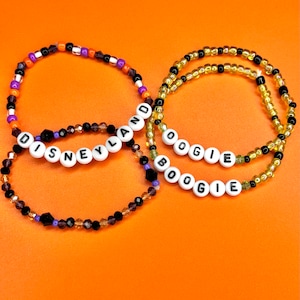 May include: Four beaded bracelets with white letter beads spelling "DISNEYLAND" and "BOOGIE". The bracelets feature black, orange, purple, and gold beads. The background is a solid orange color.