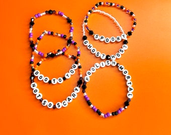 May include: Several beaded bracelets with Halloween-themed phrases and colors. The bracelets feature white letter beads spelling out phrases like "BOO TO YOU" and "NOT SO SCARY". Other bracelets say "SPOOKY", "GHOST", "FOOLISH", and "MORTAL". The beads are black, orange, purple, and clear.