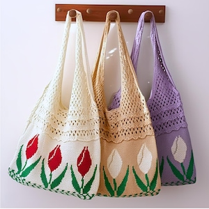 May include: Three crocheted tote bags in cream, tan, and lavender, each with a tulip design. The cream bag features red tulips, while the tan and lavender bags have white tulips. The bags hang from a wooden rack.