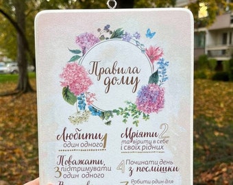 May include: A white sign with a floral design and the text "Rules of the house" in brown lettering. The sign lists eight rules for living together in a home, including "Love one another", "Dream", "Respect", "Start the day with a smile", "Fulfill your obligations", "Forgive", "Be kind", and "Say "thank you".