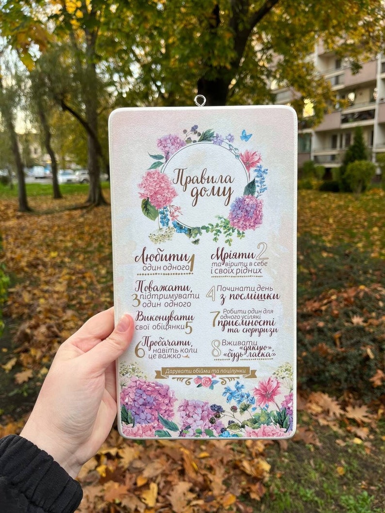 Op de afbeelding: Een wit bord met een bloemenontwerp en de tekst "Huisregels" in bruine letters. Het bord somt acht regels op voor het samenleven in een huis, waaronder "Liefhebben", "Dromen", "Respect", "Begin de dag met een glimlach", "Voldoe aan je verplichtingen", "Vergeef", "Wees aardig" en "Zeg "dank je wel".