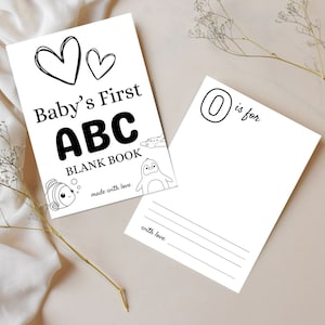 Può includere: Un libro bianco "Baby's First ABC Blank Book" con testo nero e illustrazioni di un pesce, un aereo e un pinguino. La copertina presenta due cuori. Una pagina separata mostra "O is for" e linee per scrivere.