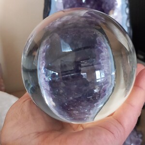 Peut inclure: Une sphère de verre transparente, également appelée boule de cristal, est tenue dans une main. La sphère reflète un cristal d'améthyste violet et blanc.
