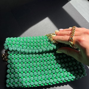 Peut inclure: Une pochette rectangulaire composée de petites perles vert vif disposées selon un motif en grille uniforme. Le sac est doté d'une bandoulière en chaîne métallique dorée et d'un fermoir assorti en métal doré sur le rabat supérieur.