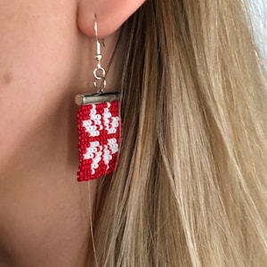 May include: A rectangular drop earring featuring a woven bead pattern in vibrant red and white. The design consists of four interconnected floral-like shapes. The beadwork is attached to a silver-toned metal finding with a standard hook wire.