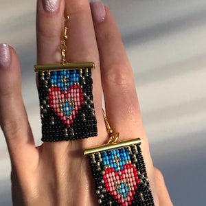May include: A pair of rectangular beaded earrings featuring a black background with a central heart design in shades of pink, red, and blue. The earrings are finished with a gold-toned metal bar at the top and gold-toned ear wire hooks for hanging.