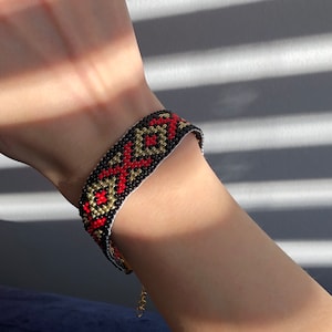 May include: A black, red, and gold beaded bracelet with a geometric pattern. The bracelet is on a person's wrist.