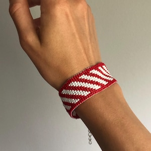 May include: A red and white beaded bracelet with a chevron pattern. The bracelet is worn on a wrist and has a silver-colored chain clasp. The bracelet's design features alternating rows of red and white beads, creating a zig-zag pattern.