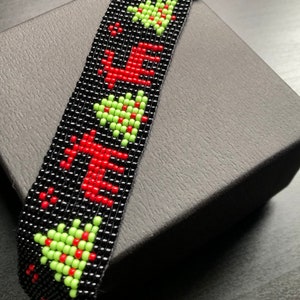 May include: A woven bead bracelet featuring a black background with a repeating pattern of red reindeer and light green Christmas trees. The bracelet is finished with silver-toned metal end bars and a circular jump ring clasp at the top edge.