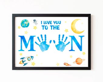 May include: Framed art print with a black border. The print features the text "I LOVE YOU TO THE MOON" in blue, with handprints replacing the Os. Space-themed illustrations include a rocket, planet, and stars.