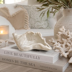 May include: A white ceramic shell-shaped bowl resting on two stacked books titled Live Beautifully and Homes by the Sea. The bowl has an irregular, organic edge and a glossy finish, positioned next to a white coral piece and a textured vase.