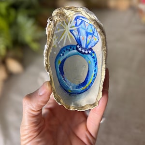 May include: Hand-painted oyster shell featuring a blue and white diamond ring design. The ring is painted with blue and yellow accents, set against a light background. The shell's edge is trimmed with gold paint.