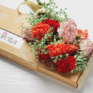 以下が含まれることがあります： 赤、オレンジ、ピンクのカーネーションと緑のカスミソウの花束。茶色のギフトボックスに入っており、ジュートのハンドルと「Loretta」の文字が入った白いラベルが付いています。