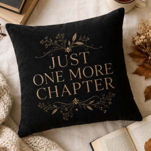 May include: A square black throw pillow featuring the gold-colored text "JUST ONE MORE CHAPTER" centered on the front. The text is framed by delicate gold botanical illustrations of leaves and sprigs above and below the lettering.
