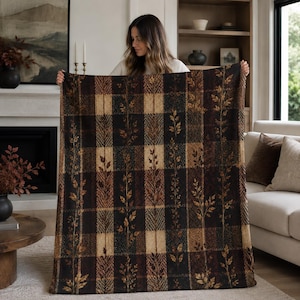 May include: A rectangular throw blanket featuring a dark brown and tan plaid pattern. The design includes vertical columns of woven leaf motifs alternating with textured, solid-colored blocks in shades of black, deep brown, and warm beige.