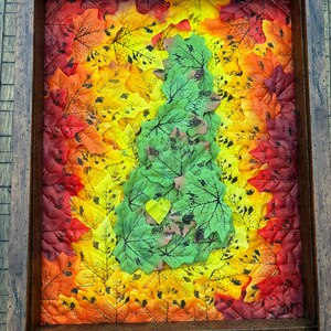 May include: A framed artwork featuring a green heart-shaped design surrounded by a gradient of fall leaves in shades of red, orange, and yellow.