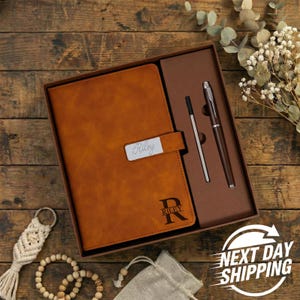 May include: A brown leather journal with the name "Riley" engraved on a silver plate. The journal is in a gift box with a pen and a stylus. The journal has the letter "R" engraved on the bottom right corner. The words "Next Day Shipping" are visible.