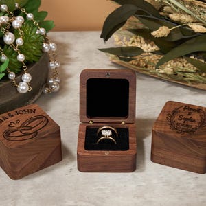 May include: Three wooden ring boxes, two closed and one open, displaying an engagement ring. The open box reveals a diamond ring set in a gold band, nestled in black velvet. The closed boxes are engraved with names and dates.