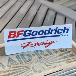May include: A white sticker with blue and red lettering that reads "BFGoodrich Tires Racing". The sticker is on a wooden surface.