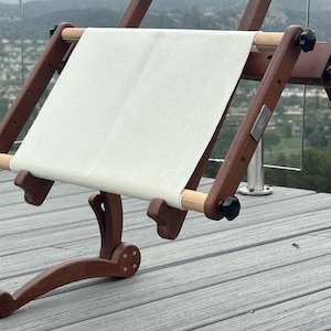 May include: A wooden embroidery stand with a white canvas stretched across it. The stand is adjustable and has a curved base. The stand is on a gray wooden deck.