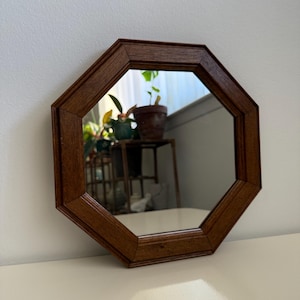 以下が含まれることがあります： 濃い茶色の木製フレームが付いた八角形の鏡。鏡は白い表面に置かれています。反射には、鉢植えの植物と金属製のスタンドが映っています。