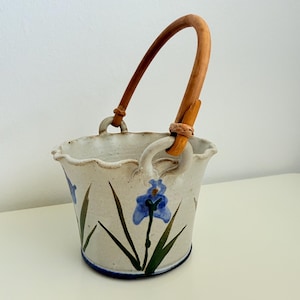May include: A ceramic bucket with a wooden handle. The off-white bucket features blue iris flower designs and green leaves. The rim has a wavy edge, and the handle is attached with leather-like material.