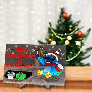 May include: A Christmas countdown sign featuring the character Stitch from Lilo & Stitch. Stitch is wearing a Santa hat and scarf. The sign has the text "Isias Countdown to Christmas" with snowflake and gift box graphics. The sign is displayed on a grey stand.