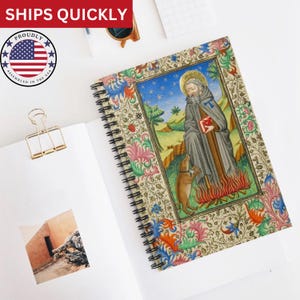 Puede incluir: Cuaderno de espiral con una ilustración detallada de un santo y un perro, sobre un paisaje con cielo azul. La portada está enmarcada con coloridos motivos florales. El cuaderno está abierto sobre una superficie blanca.