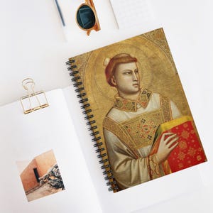 Puede incluir: Cuaderno de espiral con un retrato renacentista de un hombre con un halo dorado. La portada es una pintura detallada con detalles dorados, rojos y blancos. El cuaderno está abierto sobre una superficie blanca con un clip dorado y gafas de sol.