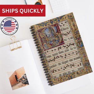 Puede incluir: Cuaderno de espiral con un diseño detallado y colorido de manuscrito medieval. La portada presenta ilustraciones y texto ornamentados, con notación musical. El cuaderno está abierto, revelando páginas en blanco y un clip dorado. El texto "SHIPS QUICKLY" es visible.