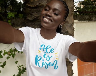 Könnte beinhalten: Ein weißes T-Shirt mit dem Text "He is Risen" in blauen und gelben Buchstaben.