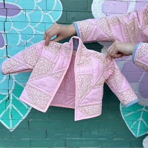May include: A light pink quilted jacket with floral patchwork details. The jacket has a light blue trim and is held up against a colorful mural. The jacket is a small size, likely for a child.