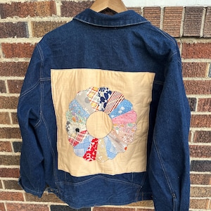 May include: A dark blue denim jacket with a beige patch on the back. The patch features a colorful, circular quilt design with various fabric patterns. The jacket has a classic collar, buttoned cuffs, and a wooden hanger.