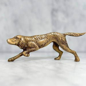 Op de afbeelding: Een messing hondenbeeldje in een rennende pose. De sculptuur heeft een getextureerd oppervlak en is afgewerkt in een warme, antieke goudkleur. De vorm van de hond is slank en atletisch, met de poten uitgestrekt in volle gang. Een decoratief item.