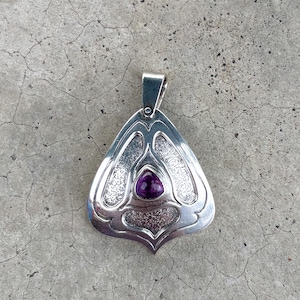 May include: A silver pendant with a triangular shape and a smooth, polished finish. The center features a small, teardrop-shaped purple gemstone. The metal surface has textured, recessed areas surrounding the stone, and a silver bail at the top.