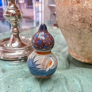 May include: A small, hand-painted gourd-shaped vase with a dark brown top dotted with blue spots. The body features a bird illustration with blue and orange accents, set against a gray background. The vase is on a green surface.