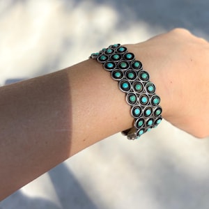 Op de afbeelding: Een zilverkleurige armband met drie rijen cirkelvormige zettingen, elk met een kleine, ronde turkoois steen. De armband wordt om een pols gedragen, waarbij het gedetailleerde ontwerp en de levendige kleur van de stenen worden getoond.