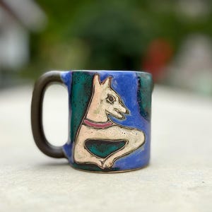 Puede incluir: Una taza de cerámica con un asa marrón oscuro. La taza presenta una figura blanca con forma de perro con un collar rosa sobre un fondo azul y verde. La taza está hecha a mano y tiene un diseño único.