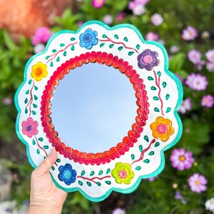 Puede incluir: Espejo redondo pintado a mano con borde festoneado. El marco blanco está adornado con flores coloridas en tonos azules, amarillos, rosas y naranjas. El borde interior es rojo y el borde exterior es turquesa.