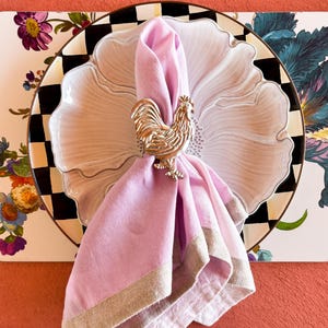May include: A decorative table setting featuring a pink linen napkin held by a gold rooster napkin ring. The napkin rests on a white floral plate with a black and white checkered rim. The background includes a floral patterned placemat.